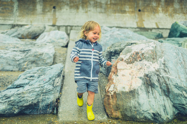 A little toddler is running and playing on a rocky beach in the rain - Fotó, kép