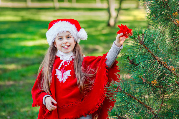 Portrait of a little girl in a Santa Claus hat near a Christmas tree in the forest. - Foto, imagen