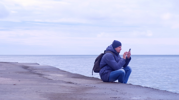 Człowiek jest wpisanie wiadomości na smartphone siedzi na morzu nabrzeżu w dzień zachmurzenie. - Materiał filmowy, wideo