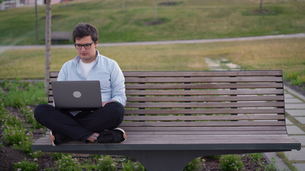 Młody człowiek korzystający z laptopa w parku siedzi na ławce na niewyraźnym tle trawy - Materiał filmowy, wideo