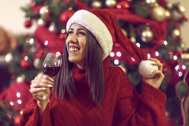 junge Frau mit Weihnachtsmütze am Weihnachtsbaum zu Hause beim Anstoßen mit Rotwein. - Foto, Bild