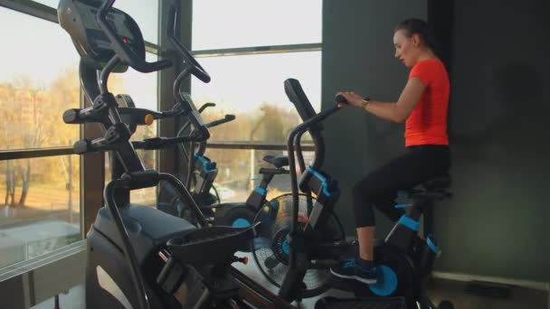 Joven mujer activa haciendo girar una bicicleta de aire en el gimnasio con los entrenadores. Entrenamiento femenino en bicicleta neumática
. - Imágenes, Vídeo