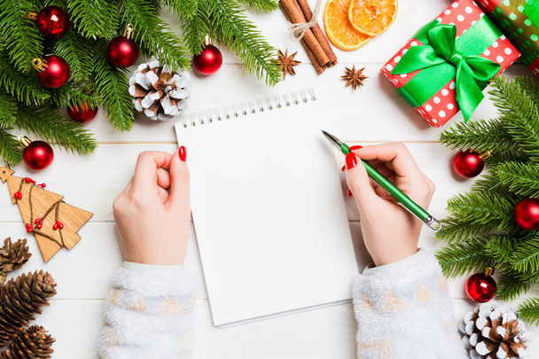 Top view of female hand writing in a notebook on wooden Christmas background. Fir tree and festive decorations. Wish list. New Year concept - Foto, Imagen