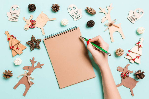 Vista superior de la mano femenina haciendo algunas notas en cuaderno sobre fondo azul. Decoraciones de Año Nuevo y juguetes. Concepto de Navidad - Foto, imagen