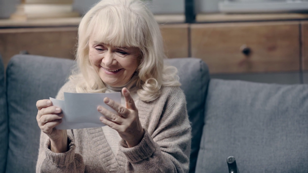 Χαμογελαστή ηλικιωμένη γυναίκα με άνοια κοιτάζοντας φωτογραφίες στον καναπέ - Πλάνα, βίντεο