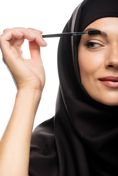 gedeeltelijk zicht van jonge moslimvrouw in hijab mascara aanbrengen op wimpers geïsoleerd op wit - Foto, afbeelding