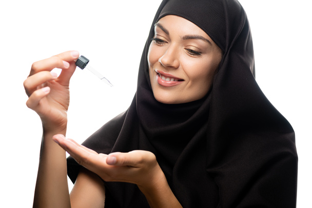smiling young Muslim woman in hijab holding dropper with serum isolated on white - Foto, afbeelding