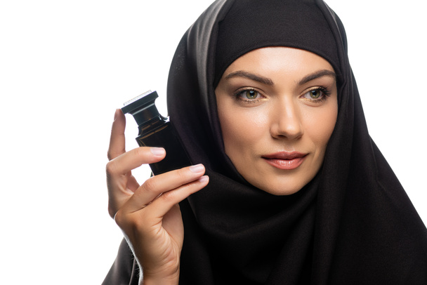 молодая мусульманка в хиджабе держит бутылку духов и смотрит в сторону изолированных на белом
 - Фото, изображение