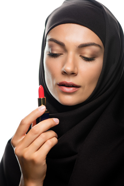 young Muslim woman in hijab looking at red lipstick isolated on white - Foto, imagen