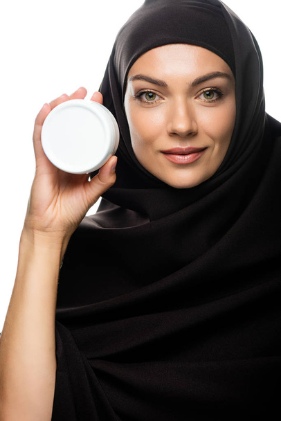 sonriente joven musulmana en hijab sosteniendo contenedor con crema cosmética aislada en blanco
 - Foto, Imagen