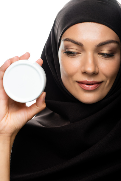 Улыбающаяся мусульманка в хиджабе держит контейнер с косметическим кремом, изолированным на белом
 - Фото, изображение