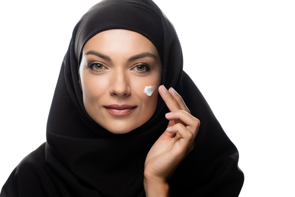 jong moslim vrouw in hijab toepassing van cosmetische crème op kuiken geïsoleerd op wit - Foto, afbeelding