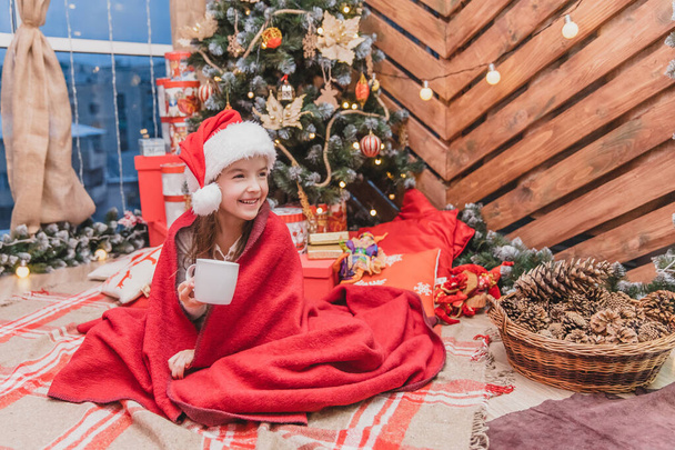 Beautiful child in santa hat, wrapped in red blanket near christmas tree, with a cup of cocoa, looking delighted and pleased. - Foto, imagen