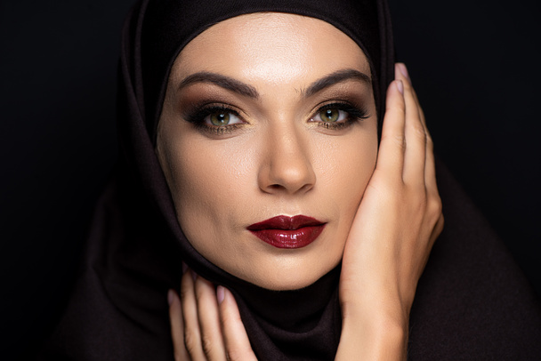jonge moslim vrouw in hijab met rokerige ogen en rode lippen geïsoleerd op zwart - Foto, afbeelding