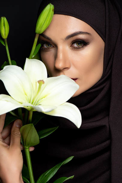 schöne muslimische Frau im Hijab mit rauchigen Augen hält Lilie isoliert auf schwarz - Foto, Bild