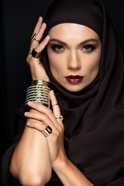 όμορφη μουσουλμάνα γυναίκα σε μαντίλα με μακιγιάζ σε χρυσά δαχτυλίδια και βραχιόλι που απομονώνονται σε μαύρο - Φωτογραφία, εικόνα