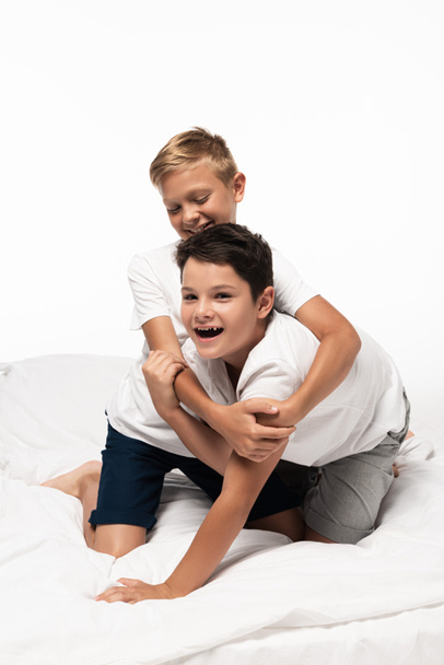 cheerful boy hugging smiling brother while having fun on bed isolated on white - Zdjęcie, obraz