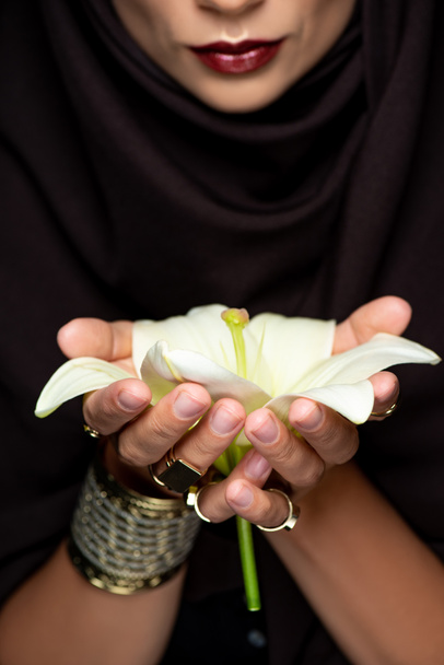 piękna muzułmanka w hidżabie w złotych pierścieniach i bransoletce trzymając lilię odizolowaną na czarno - Zdjęcie, obraz