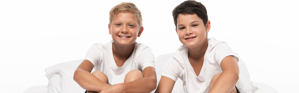 panoramic shot of two cheerful brothers sitting on bed and looking at camera isolated on white - Foto, afbeelding