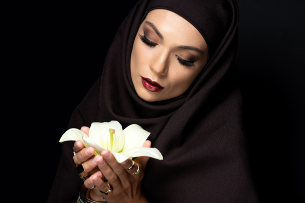 schöne muslimische Frau im Hijab in goldenen Ringen und Armband hält Lilie isoliert auf schwarz - Foto, Bild