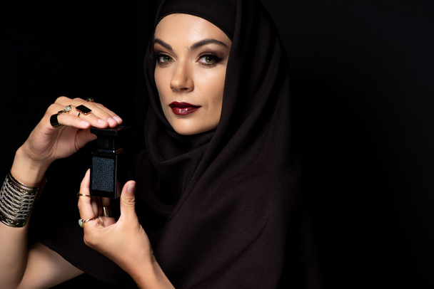 beautiful Muslim woman in hijab with makeup in golden jewelry holding perfume isolated on black - Photo, Image