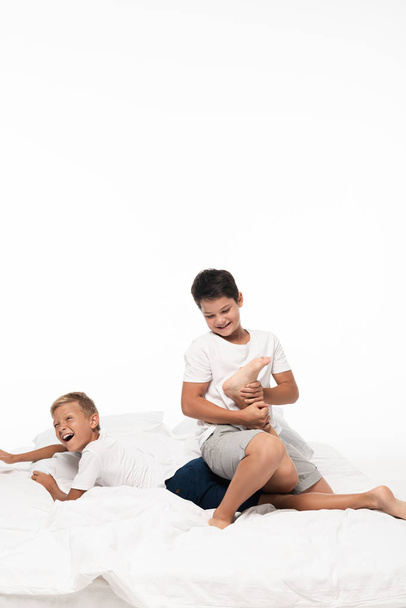 cheerful boy jokingly breaking leg of screaming brother while having fun on bed isolated on white - Photo, Image