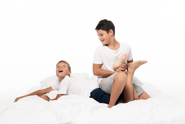 χαρούμενο αγόρι που κρατάει το πόδι των αδελφών ενώ διασκεδάζει στο κρεβάτι απομονωμένο στο λευκό - Φωτογραφία, εικόνα