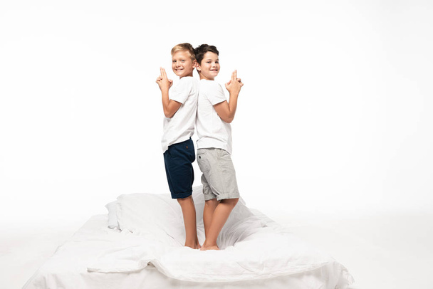 two cheerful brothers standing back to back on bed and showing gun gestures isolated on white - Fotoğraf, Görsel
