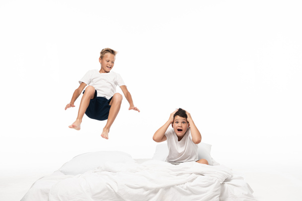 cheerful boy levitating over shocked brother sitting on bedding and holding hands on head isolated on white - Foto, afbeelding