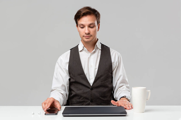 Ładny pracownik biurowy w białej koszulce i szarej kamizelce, porządkujący sprawy w swojej strefie pracy. Laptop, smartfon i filiżanka kawy na białym stole. - Zdjęcie, obraz