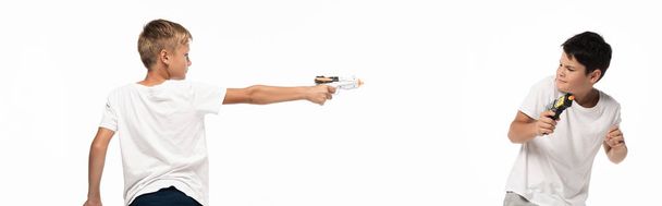 панорамный снимок мальчика, прицеливающегося игрушечным пистолетом в напуганного брата, изолированного на белом
 - Фото, изображение