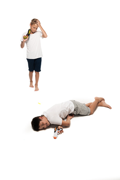 boy pretending dead near upset brother holding toy gun on white background - Photo, Image