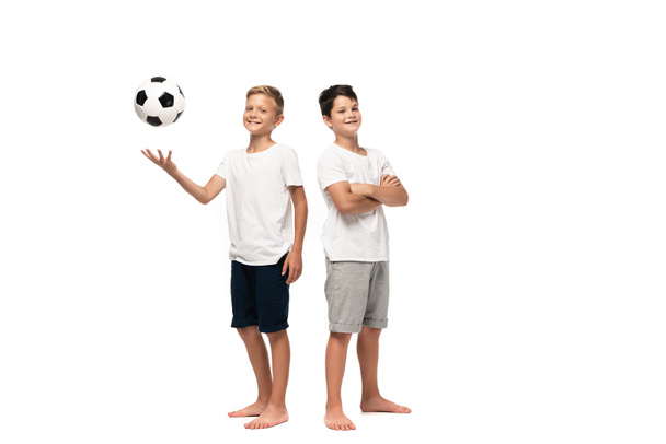 cheerful boy playing soccer ball near smiling brother standing with crossed arms on white background - Photo, Image