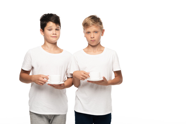 two sceptical brothers looking at camera while holding coffee cups on saucers isolated on white - Photo, image