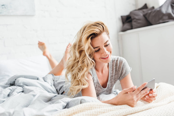 selective focus of cheerful girl using smartphone while lying on bed  - Fotó, kép