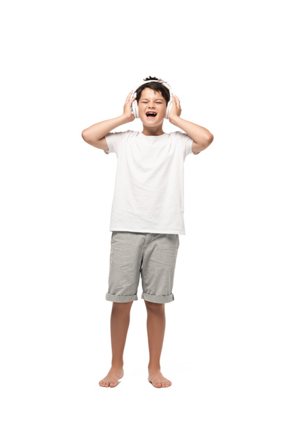 excited boy holding hands on headphones and singing while listening music on white background - Fotoğraf, Görsel