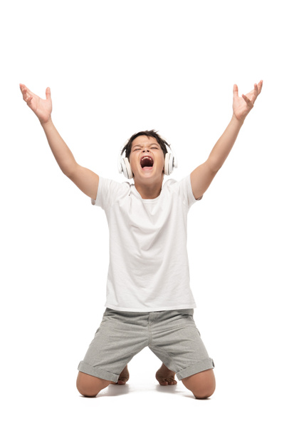 ενθουσιασμένο αγόρι στέκεται στα γόνατα με υψωμένα χέρια ακούγοντας μουσική στα ακουστικά και τραγουδώντας σε λευκό φόντο - Φωτογραφία, εικόνα
