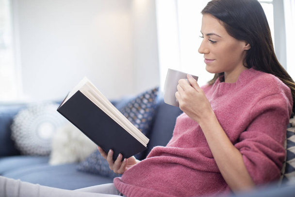 Woman With Hot Drink Relaxing On Sofa At Home Reading Book - Фото, зображення