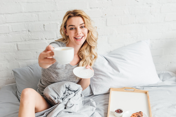 Selektiver Fokus der glücklichen Frau mit Tasse Kaffee und Untertasse im Bett  - Foto, Bild
