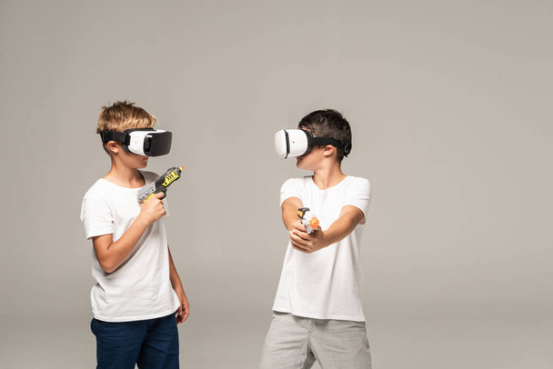 two brothers in vr headsets looking at each other while holding toy guns isolated on grey - Φωτογραφία, εικόνα