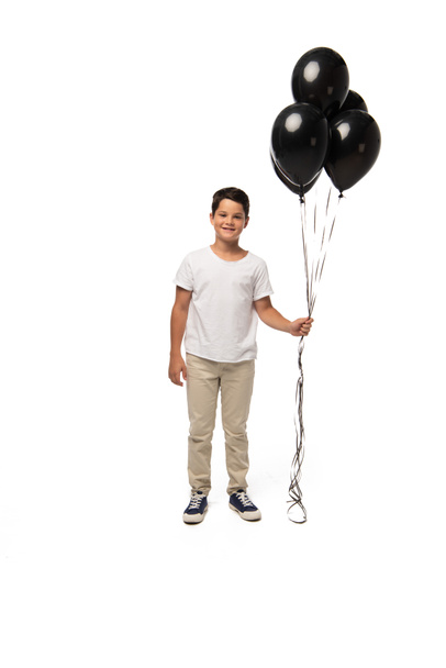 cheerful boy looking at camera while holding black balloons on white background - Valokuva, kuva