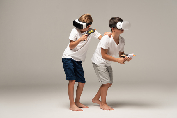 two barefoot brothers in pajamas holding toy guns while using vr headsets on grey background - Photo, Image