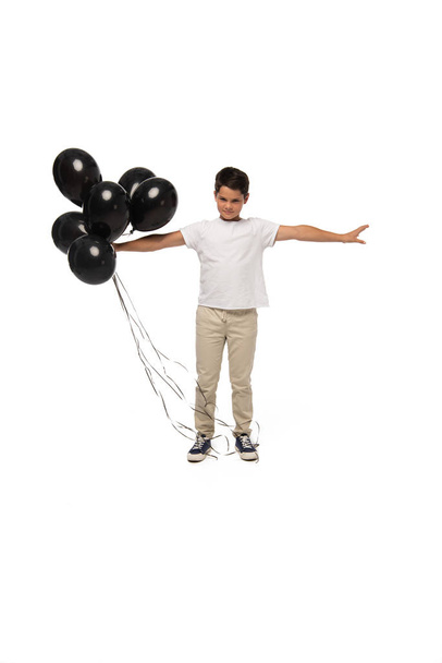 vakava poika seisoo ojennetuin käsin pitäen mustia ilmapalloja valkoisella taustalla
 - Valokuva, kuva