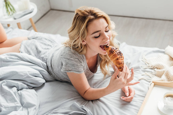 nad głową widok szczęśliwej dziewczyny jedzącej rogaliki w pobliżu tacy z filiżanką kawy na łóżku  - Zdjęcie, obraz