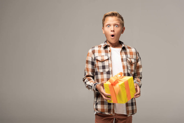 Schockierter Junge mit gelbem Geschenkkarton und Blick in Kamera isoliert auf grau - Foto, Bild