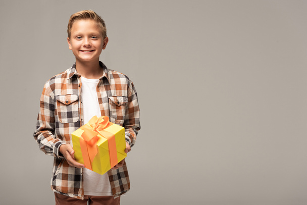 glücklicher Junge mit gelbem Geschenkkarton und lächelnd in die Kamera, isoliert auf grau - Foto, Bild