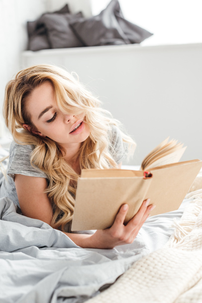 selective focus of young and attractive woman reading book while lying on bed  - Fotoğraf, Görsel