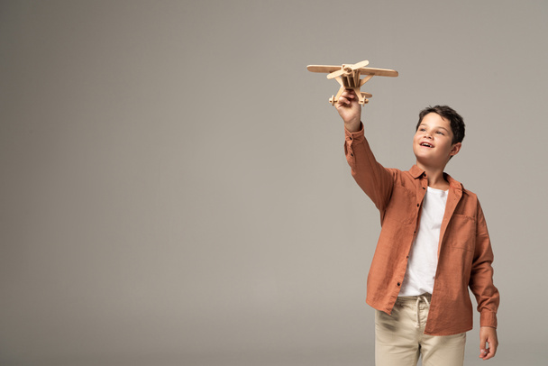 glücklicher Junge mit Holzspielzeugflugzeug in erhobener Hand isoliert auf grau - Foto, Bild