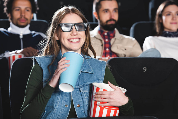 selektywne skupienie uśmiechniętej kobiety w okularach 3D trzymającej papierowy kubek i popcorn, oglądającej film w kinie  - Zdjęcie, obraz