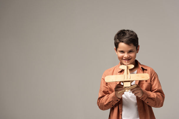 cheerful boy showing wooden toy plane and smiling at camera isolated on grey - Fotó, kép